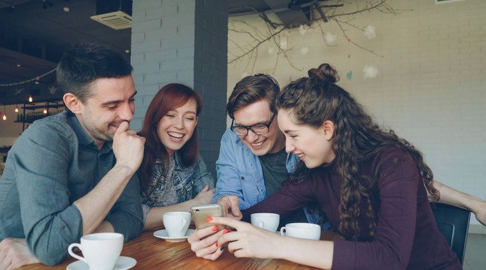 Four friends sitting at a café table, smiling and looking at a smartphone together with coffee cups in front of them.